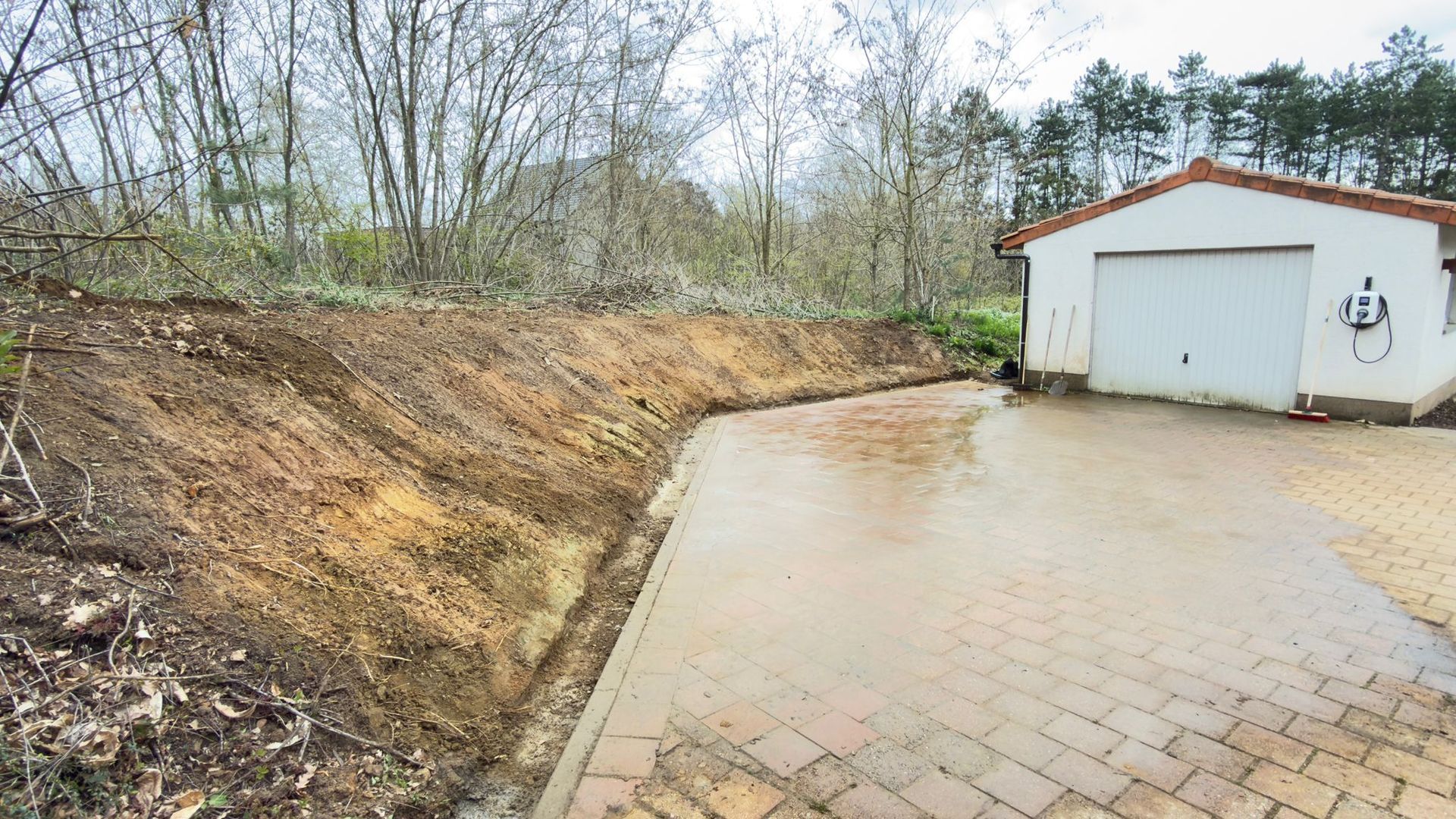 Een onverharde, met gras begroeide helling loopt omhoog naast een verharde oprit en een witte garage onder een bewolkte hemel.