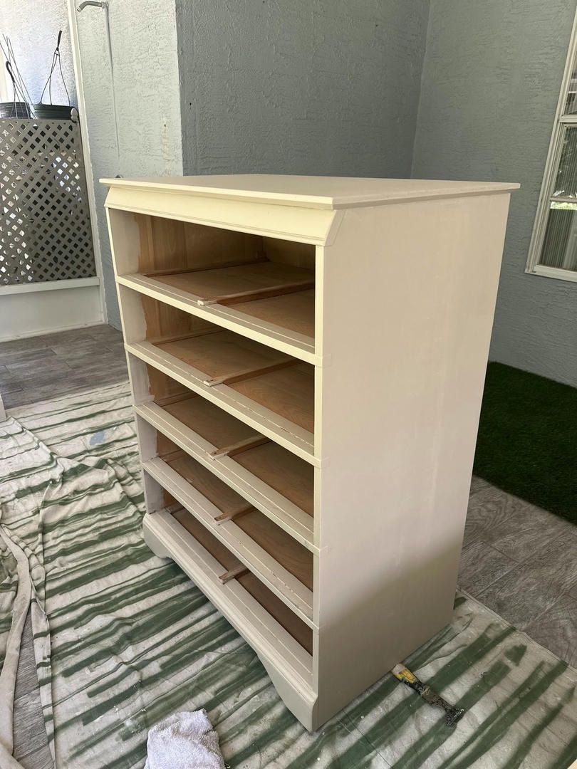 A light-colored dresser with empty drawers stands on a striped drop cloth outdoors near a wall.