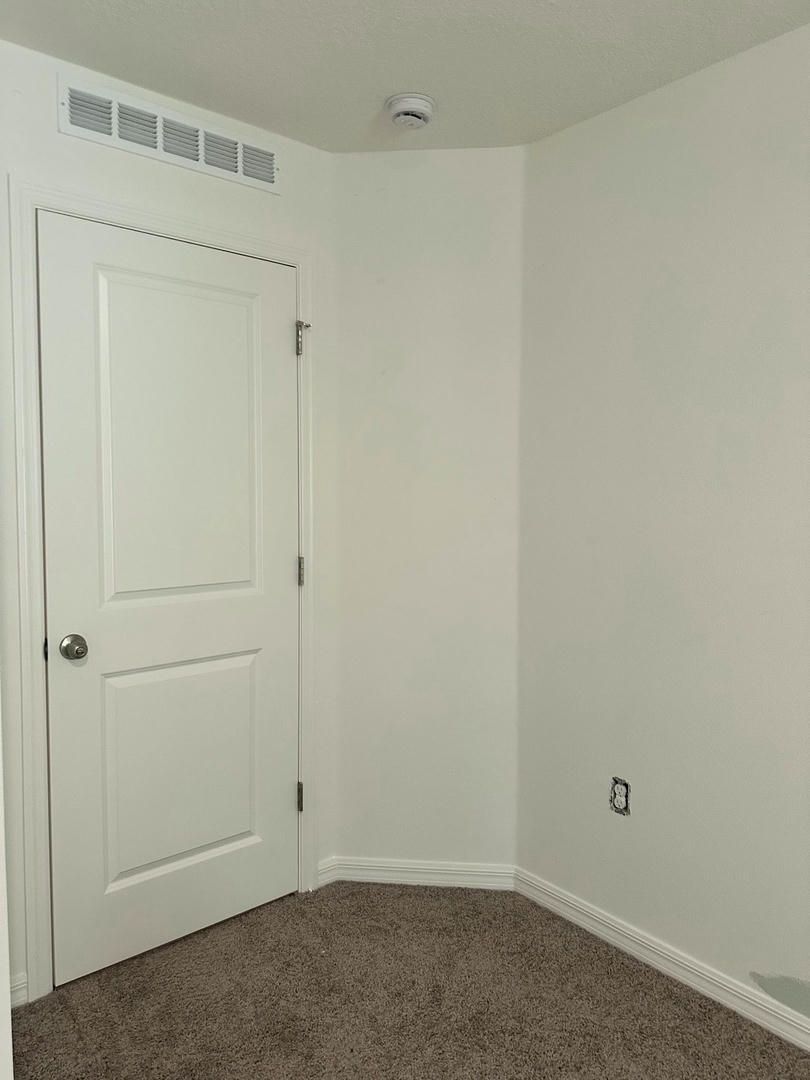 White room corner with door, vent, and electrical outlet. Tan carpet, white walls, and a smoke detector.
