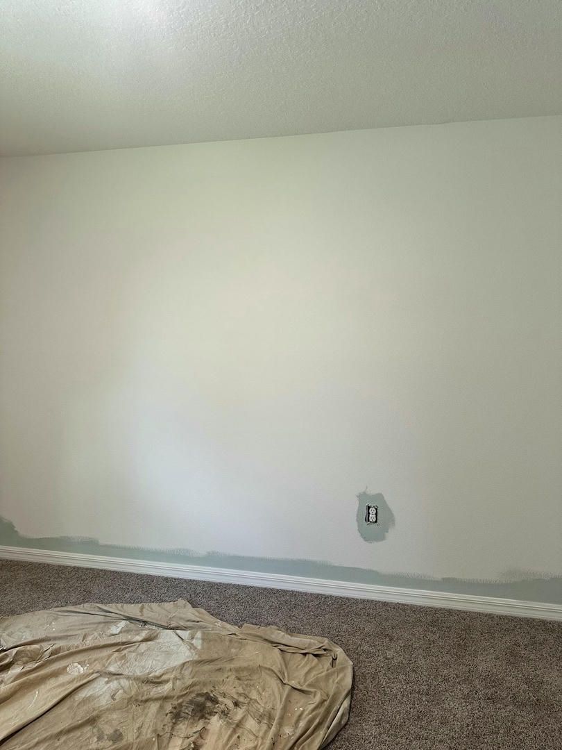 Interior wall with paint splotches and baseboard tape. Tan carpet and drop cloth on floor.