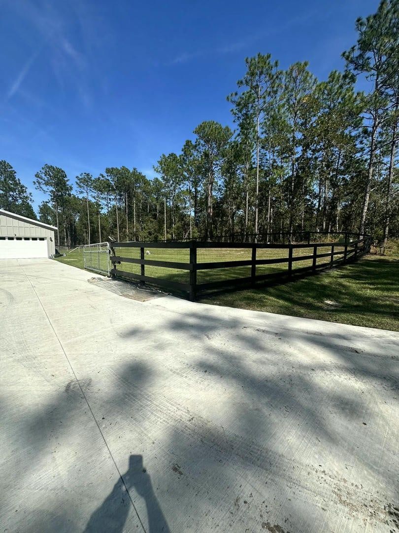 A driveway leads to a fenced-in grassy area next to a building and trees under a blue sky.