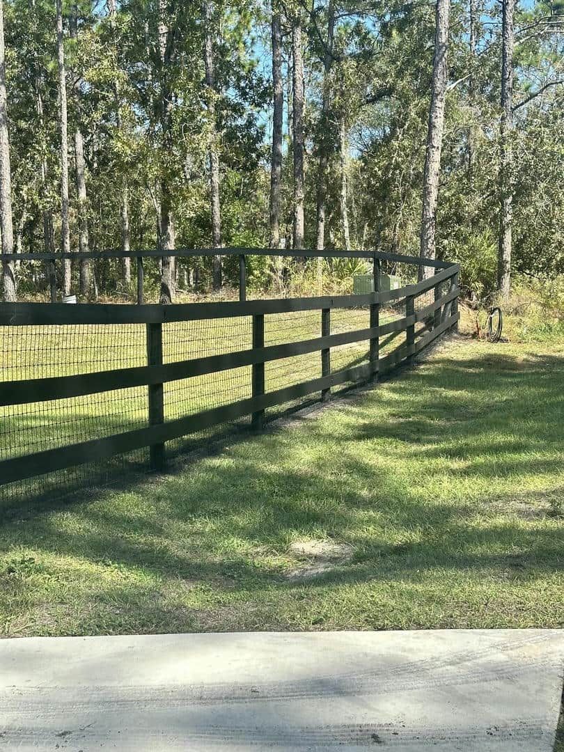 Black wooden fence in a grassy yard, trees in the background, sunny day.