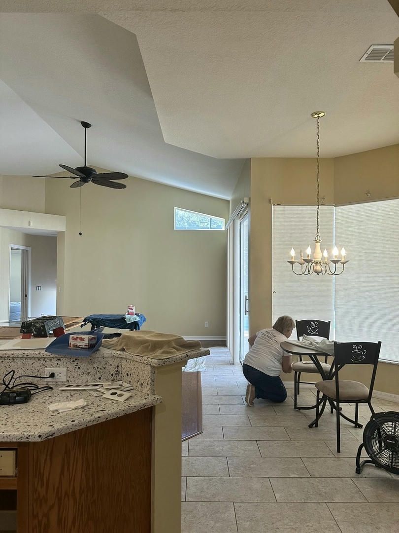 Person works at a table in a dining area; kitchen island in foreground.  Light yellow walls, tile floor, chandelier.