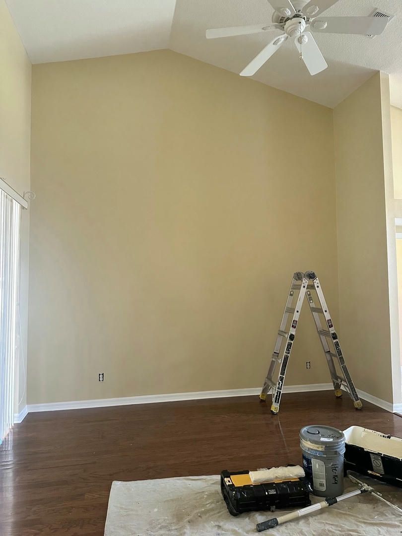 A tall beige wall with a stepladder, painting supplies, and ceiling fan in a room with wood flooring.