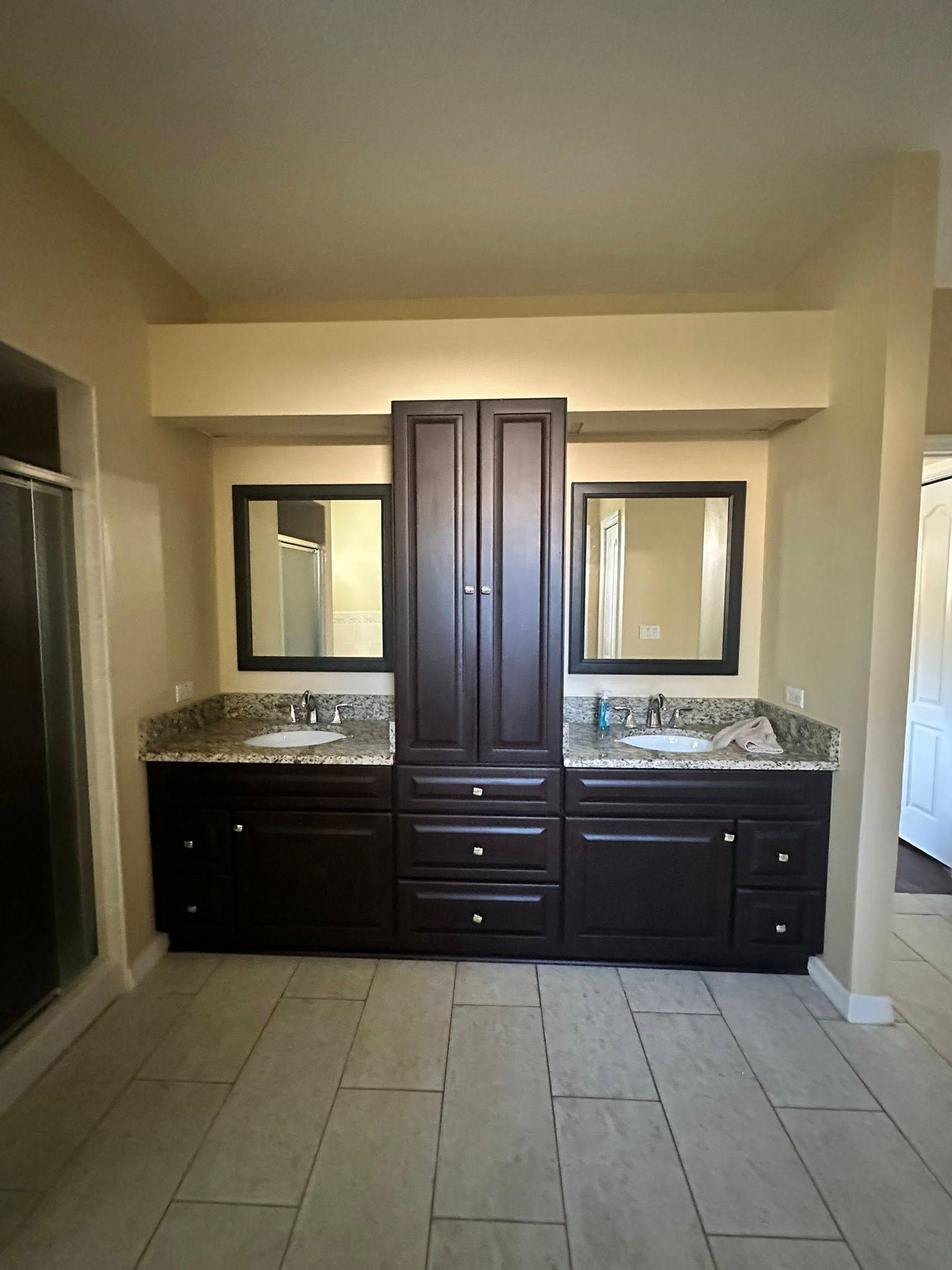 Bathroom with a dark brown double vanity, granite countertops, and a central cabinet. Two mirrors hang above the sinks.
