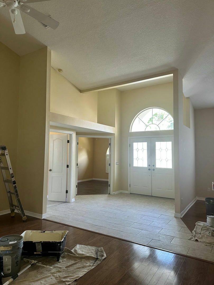 Interior of a house, featuring an entryway with high ceilings, doors, and a staircase. Painted beige and white.