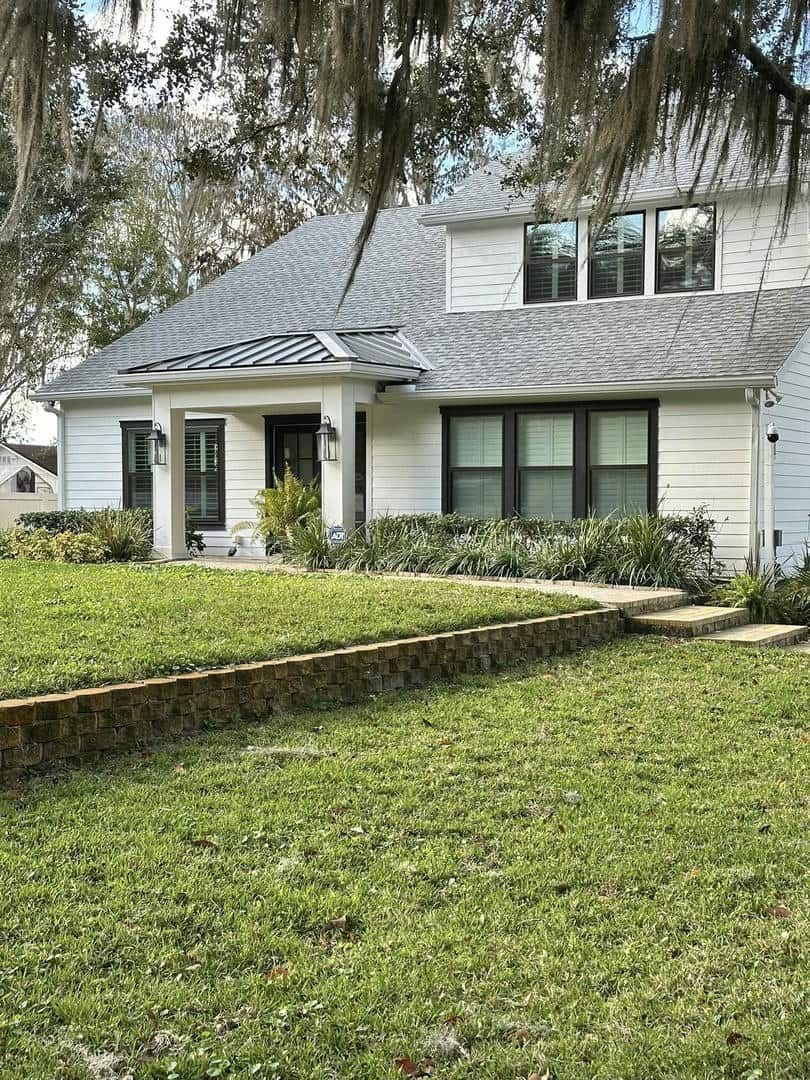 White house with a dark roof and black window frames, front yard with a low brick wall and overgrown lawn.
