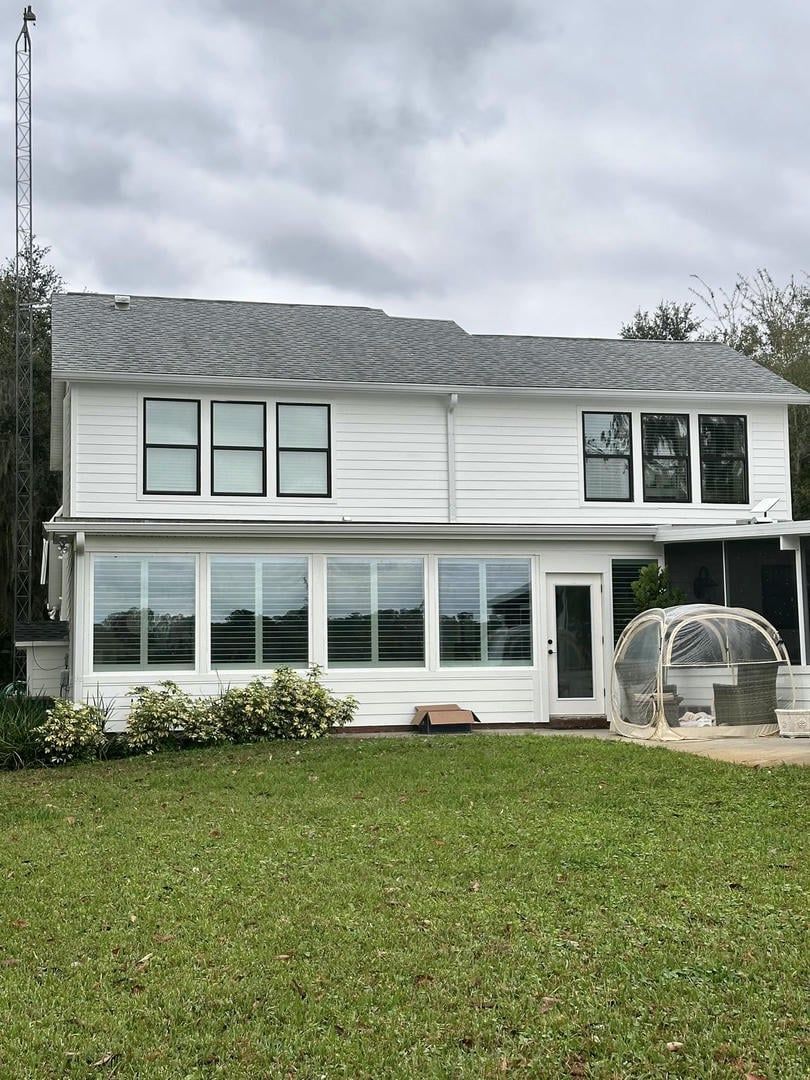 White two-story house with many windows, on a grassy lawn. Greenhouse and communication tower in view. Overcast sky.
