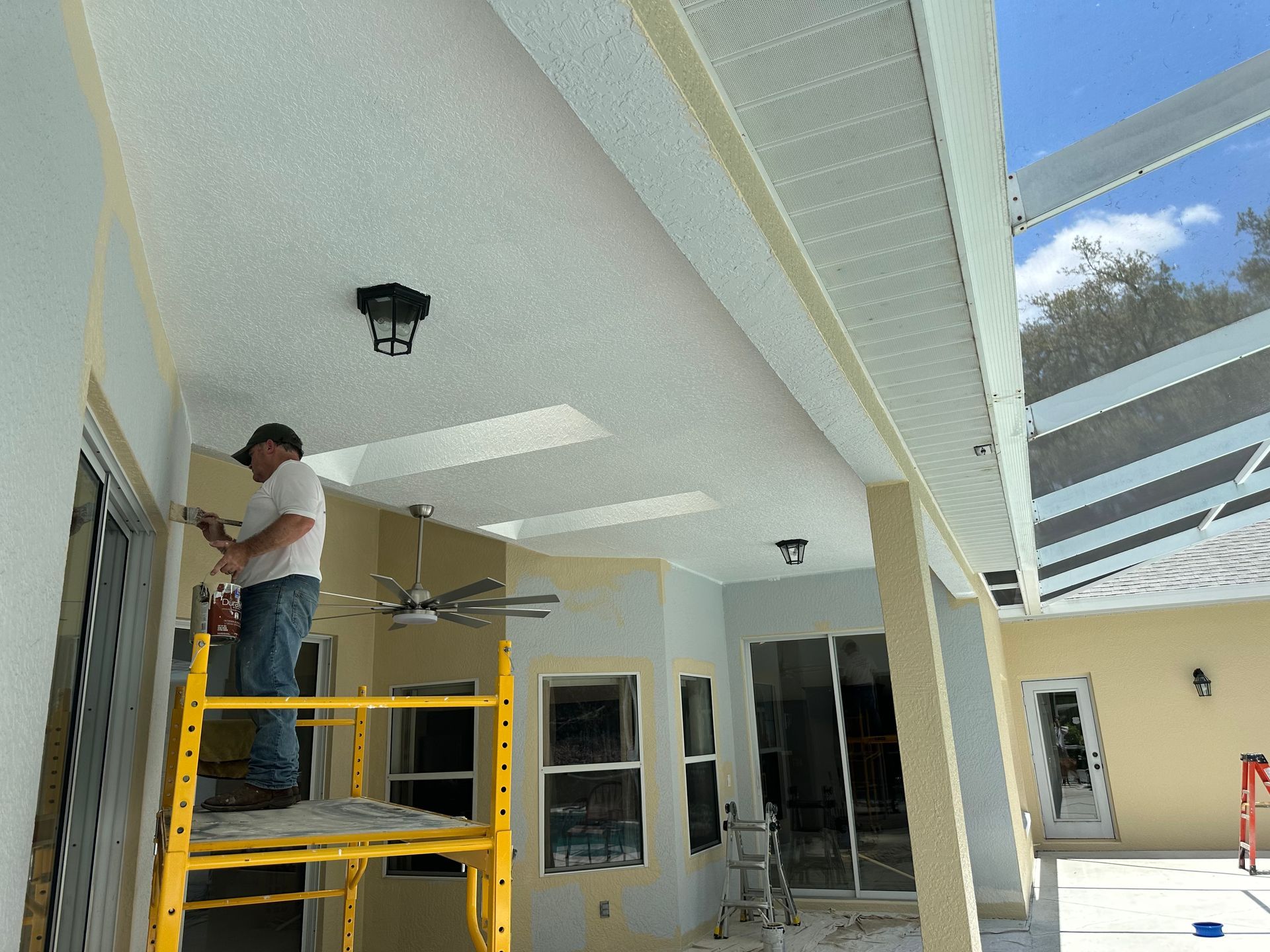 Person painting a house's exterior; white and gray colors; standing on a wooden scaffold.