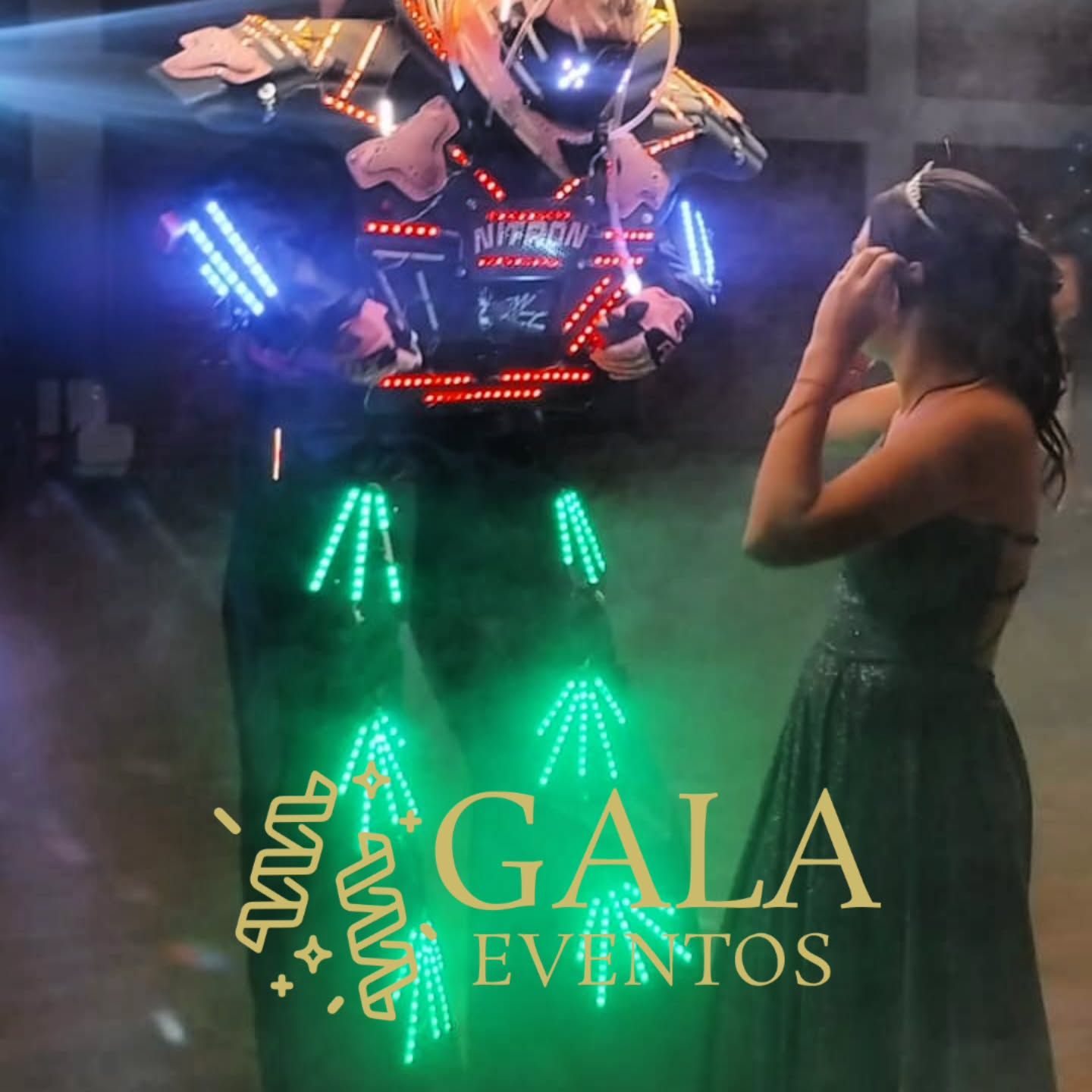 Una persona con un disfraz de robot brillante está junto a una mujer con vestido en un evento. Logotipo de