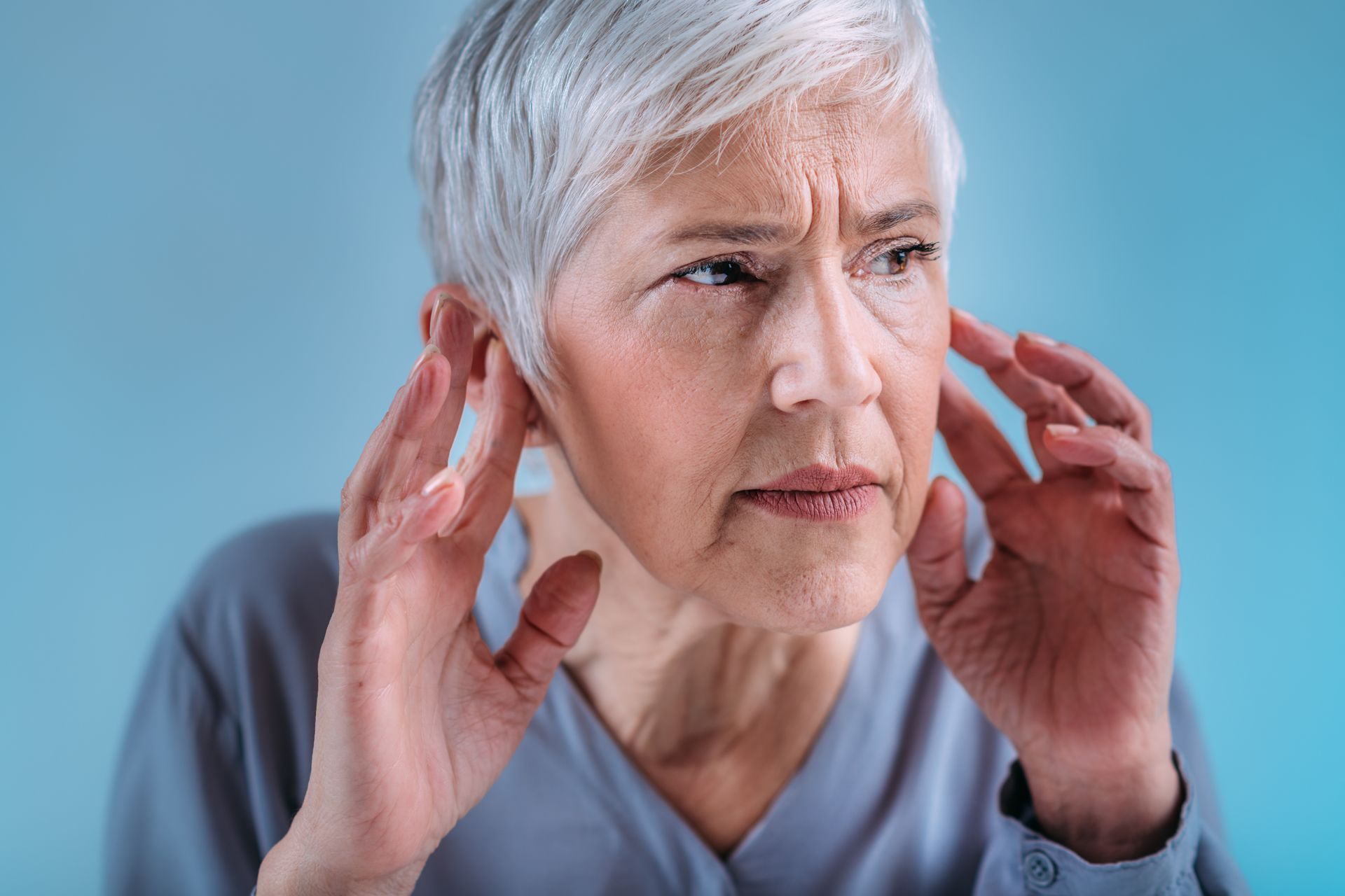 An elderly woman is covering her ears with her hands.