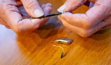 A person is fixing a hearing aid on a wooden table.