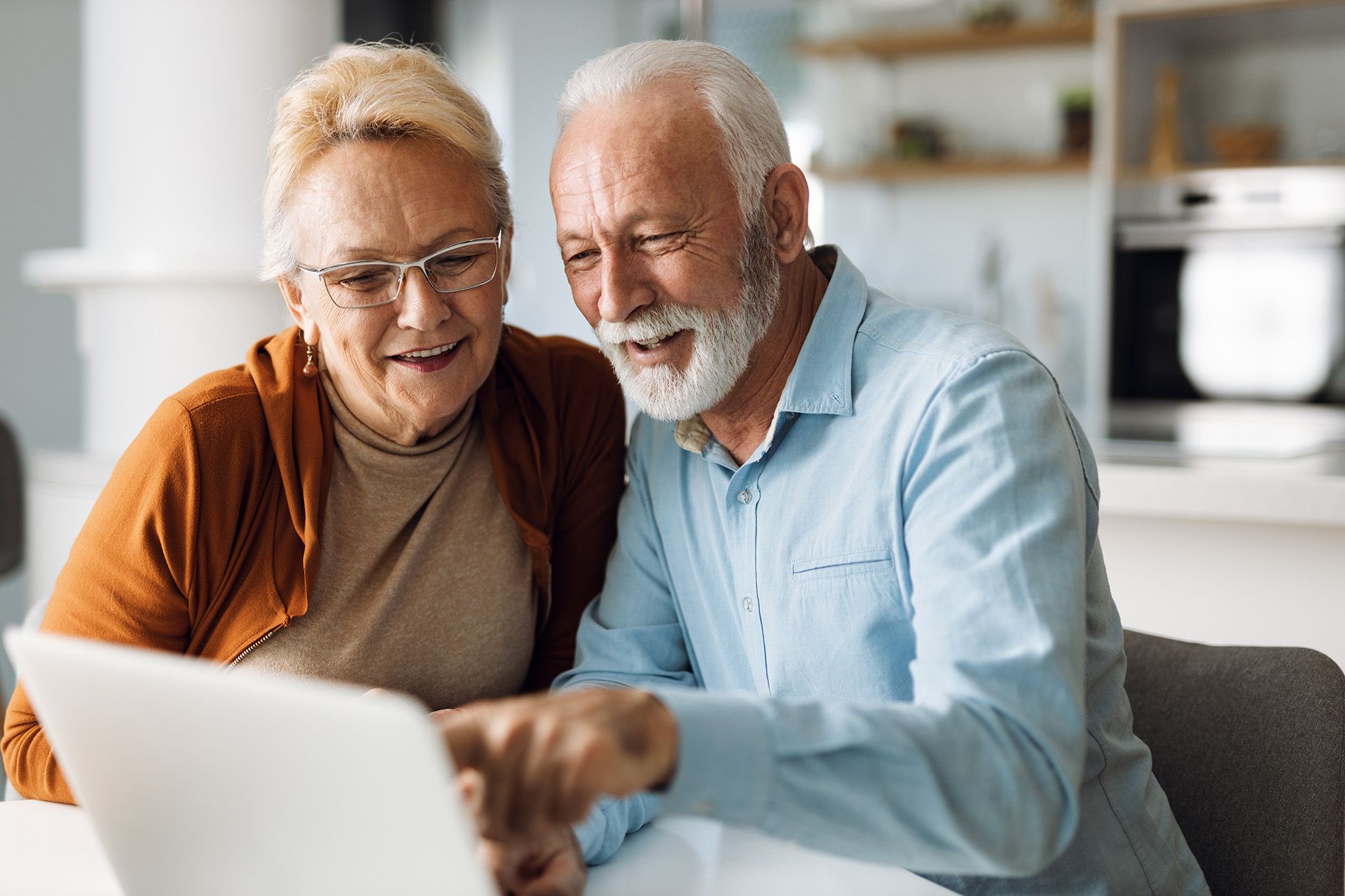 An elderly couple is sitting at a table looking at a laptop computer.