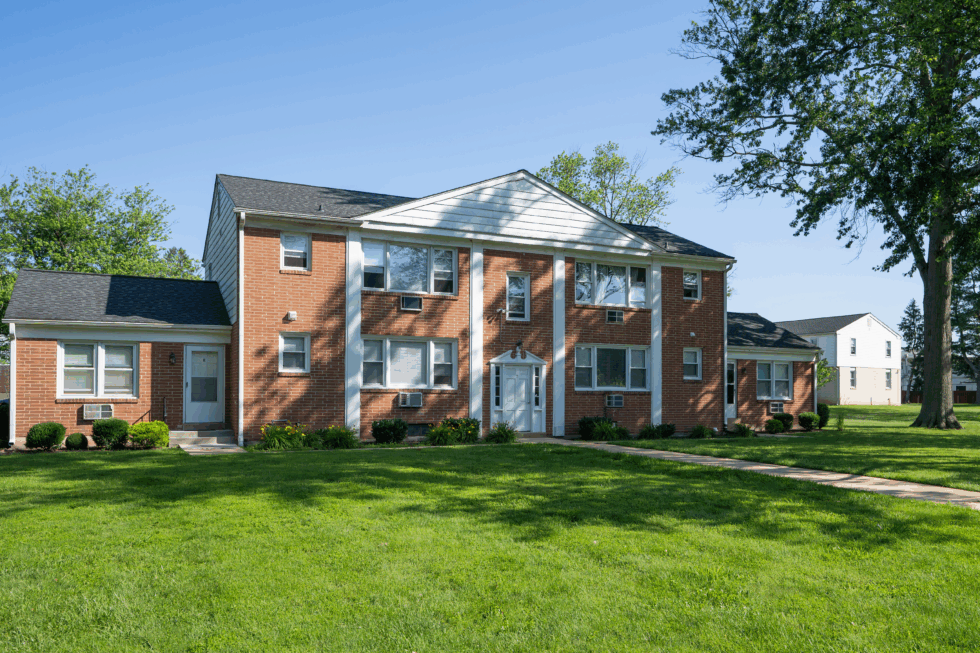 A two-story brick apartment building with a white front entrance and dark roof, set on a green lawn under a blue sky.