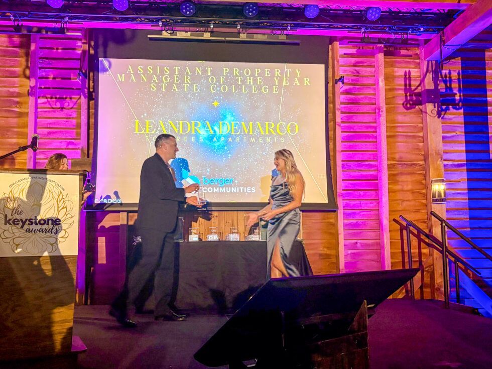 A man in a suit presenting an award to a woman in a gown on a stage with a brightly lit screen in a wood-paneled room.