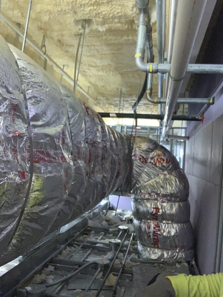 Insulated HVAC ductwork, silver, turning corner near ceiling in unfinished construction site.