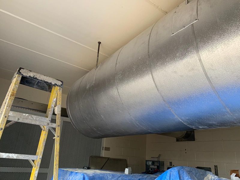 Silver-wrapped HVAC ducting suspended from a white ceiling, ladder in foreground, blue tarp below.