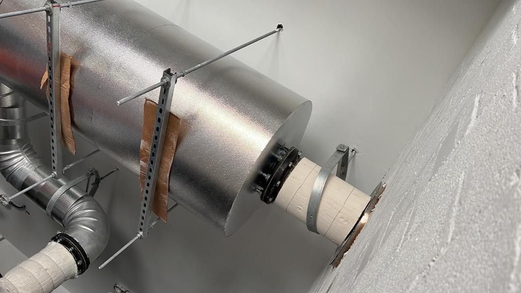 Insulated metal ductwork with support brackets, connecting through a white wall.