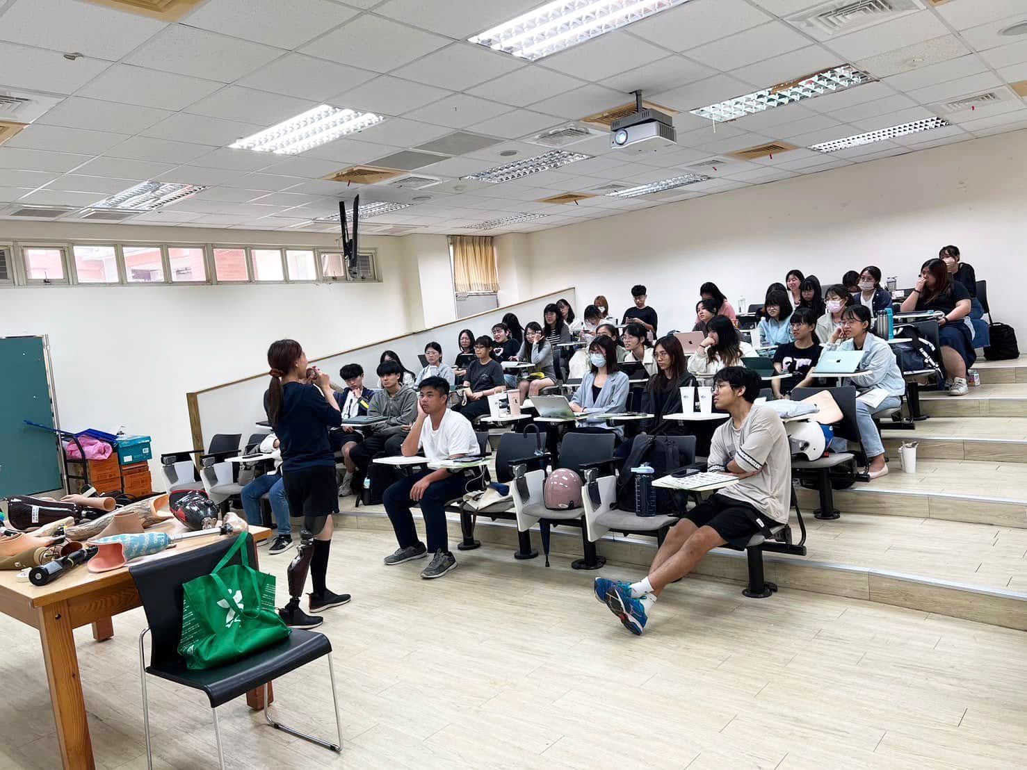 大眾教育講座現場：一名穿戴義肢的專業講師在階梯教室與學生分享，桌面上擺放多款義肢零組件，推動多元包容的友善環境。