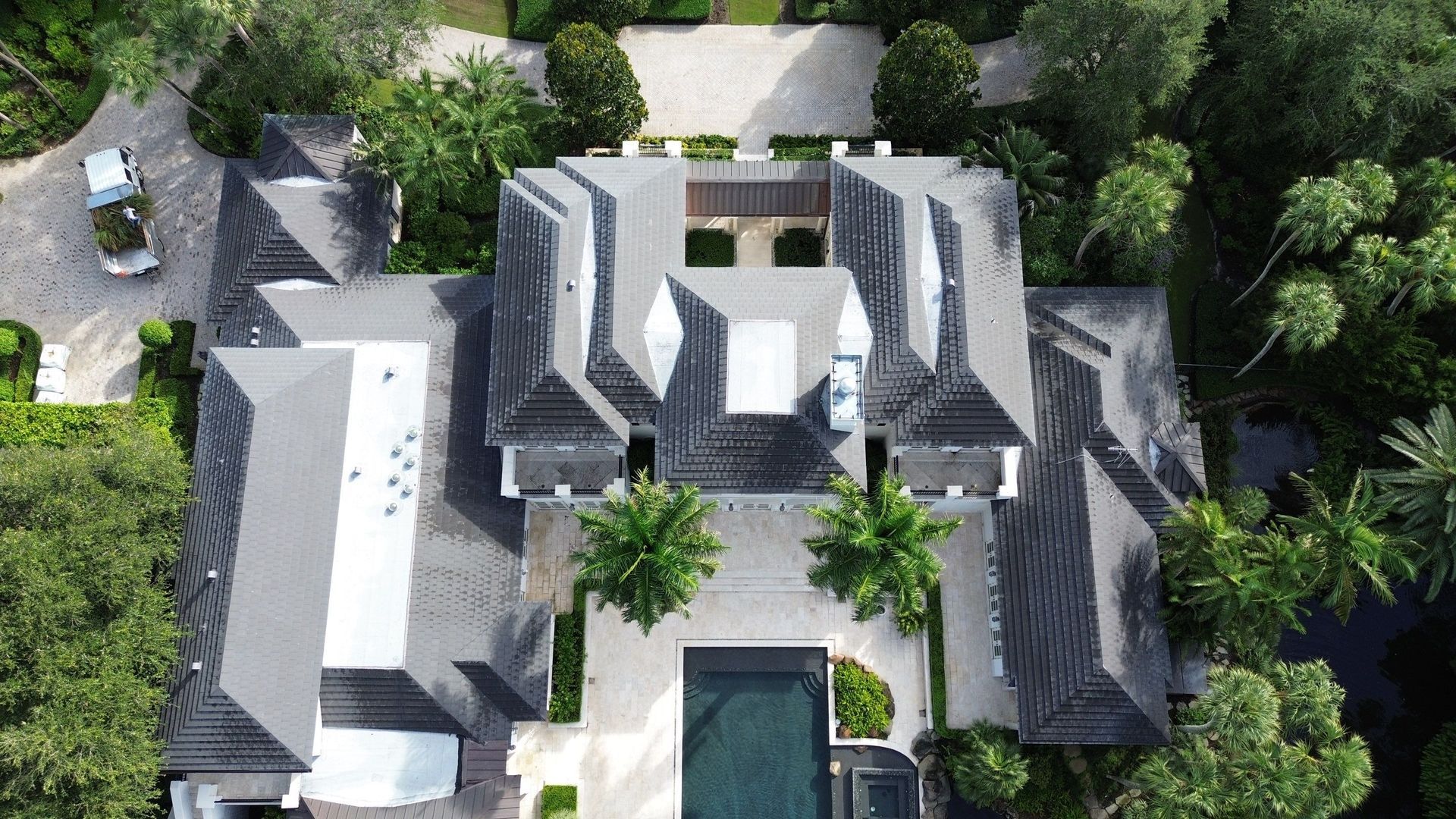 Aerial view of a large, luxury home with a dark roof, swimming pool, and lush landscaping. A white vehicle is parked in the driveway.