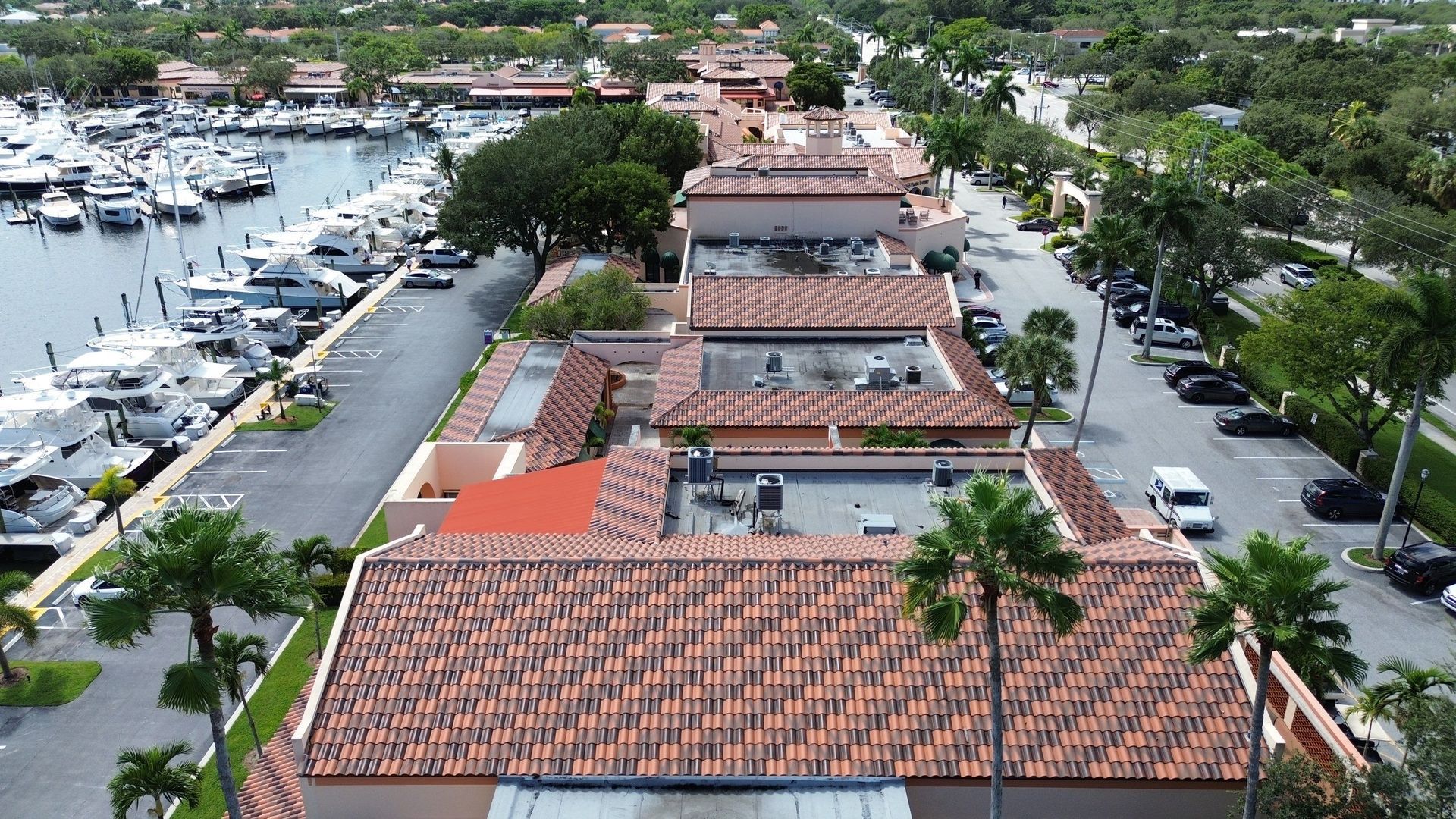 Aerial view of a building with a red-tiled roof, a marina with many boats, and a road with parked cars and palm trees.