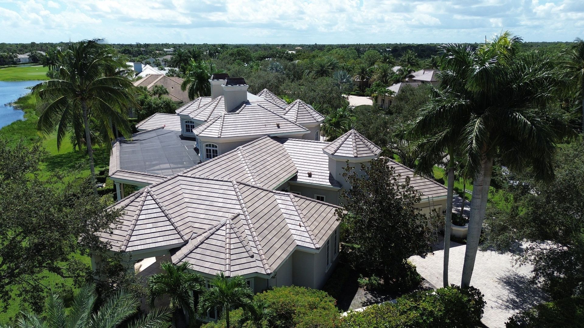 Aerial view of a large house with a light-colored roof surrounded by trees and foliage, near a lake.