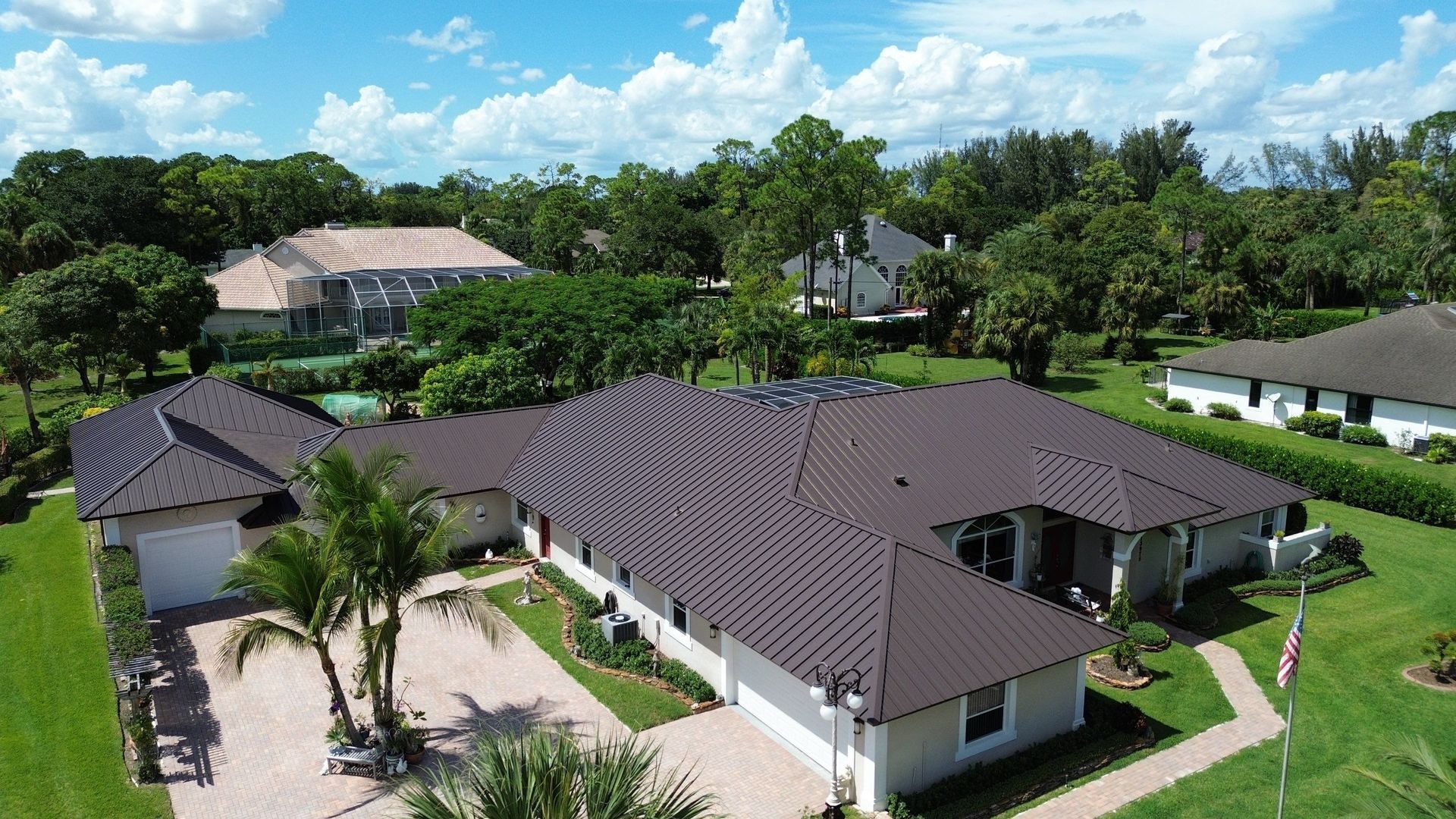 Aerial view of a beige house with a brown roof and attached garage, surrounded by green grass and trees under a blue sky.