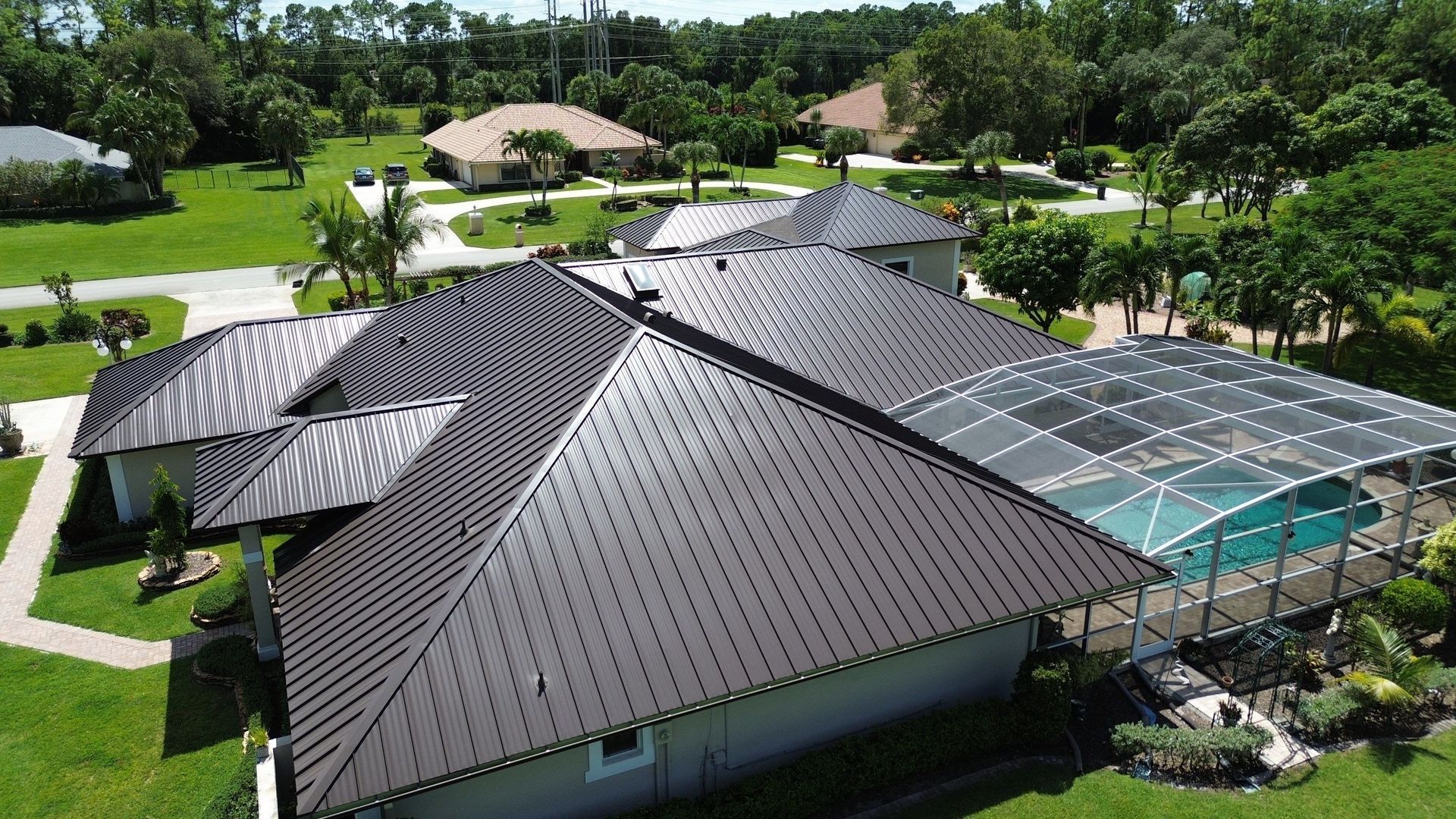 An aerial view of a house with a dark brown metal roof, green lawn, and a screened-in pool enclosure.