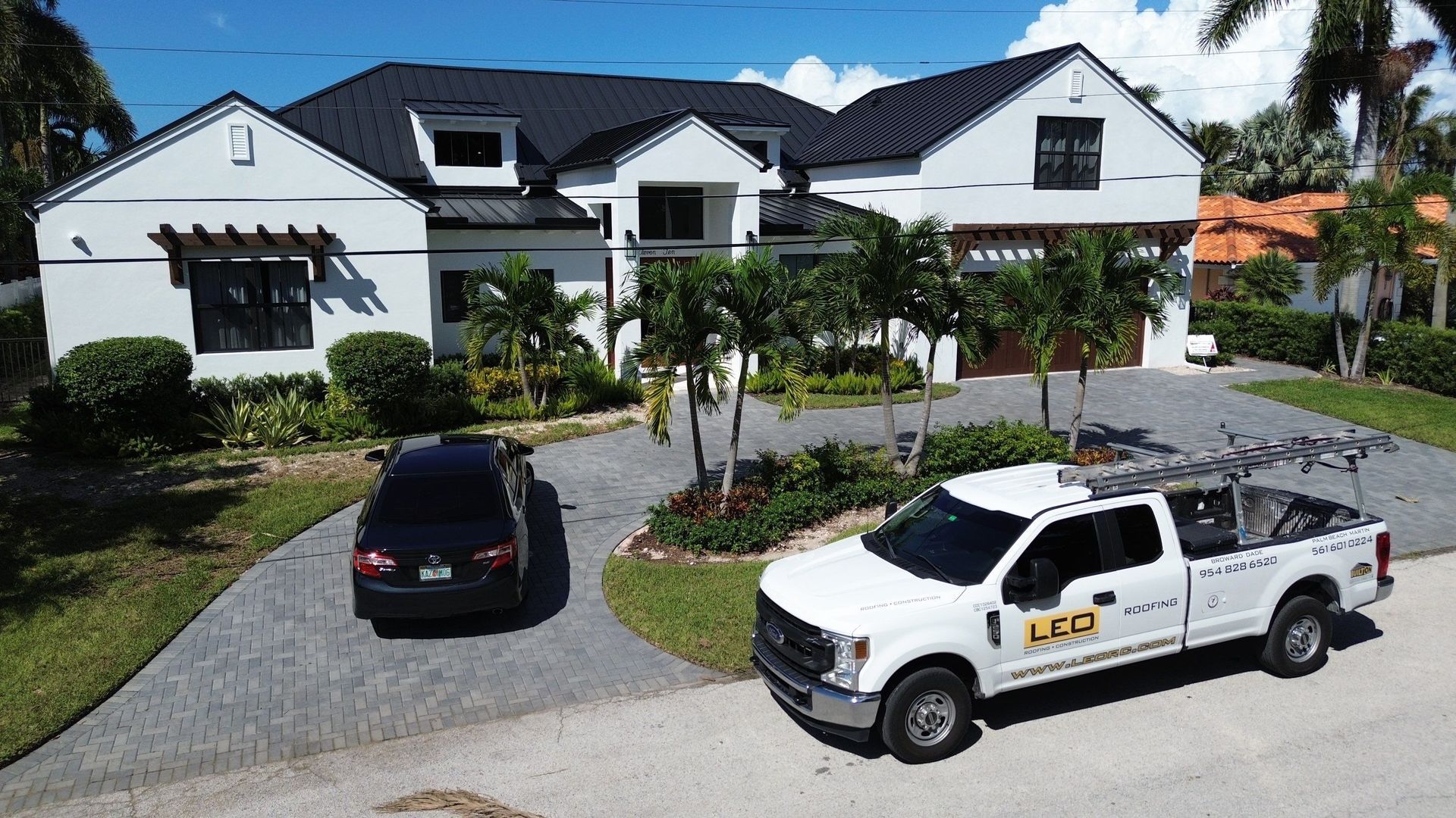 A white pickup truck and black car parked in front of a white house with a black roof on a sunny day.