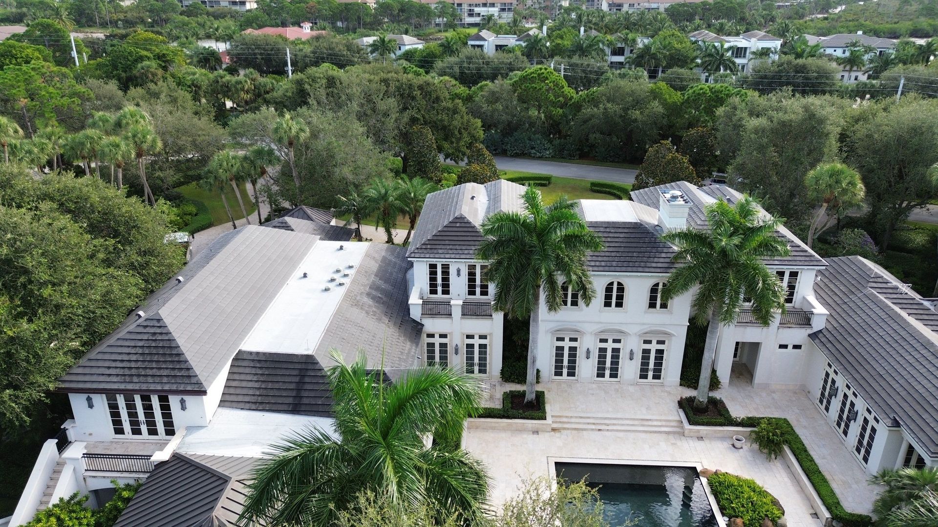 Aerial view of a large white mansion with a dark gray roof, swimming pool, and surrounding green trees.