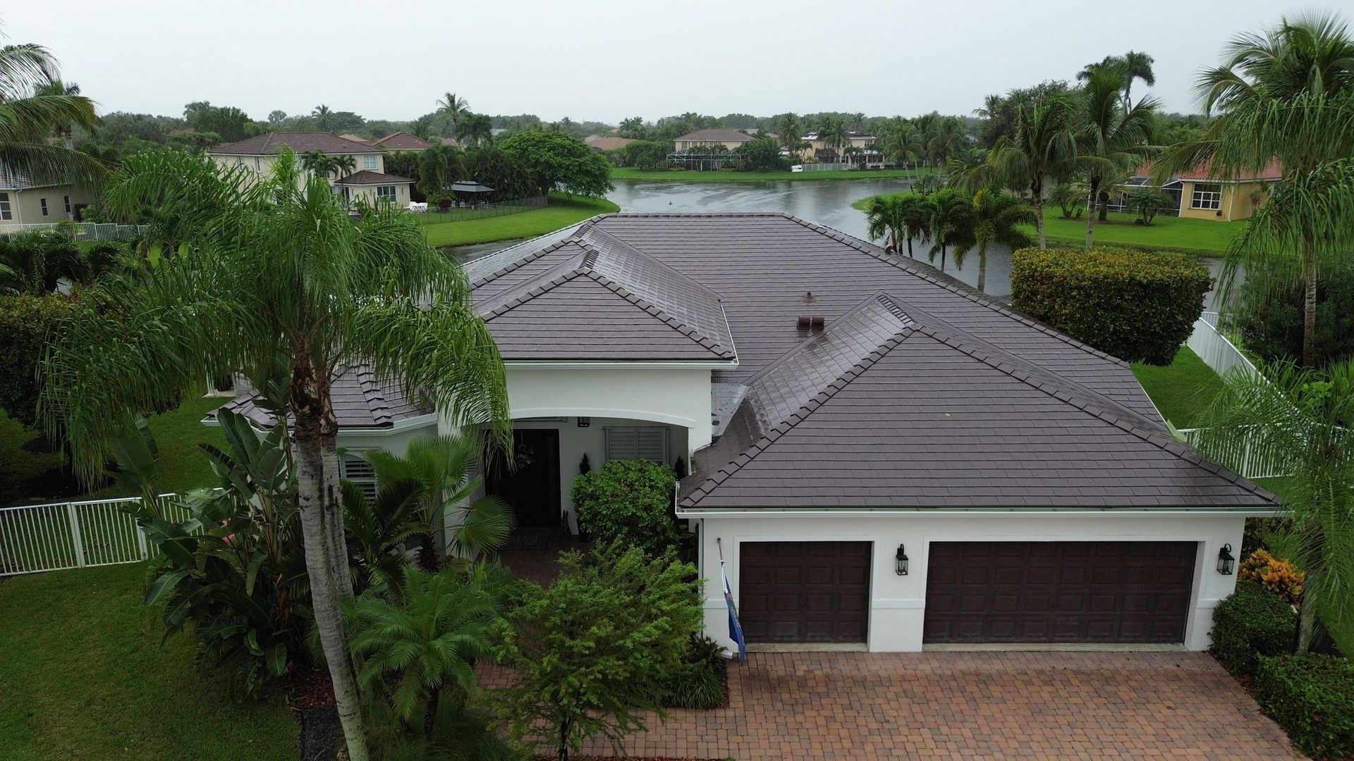 A white house with a brown roof and garage doors sits near a body of water, surrounded by greenery.