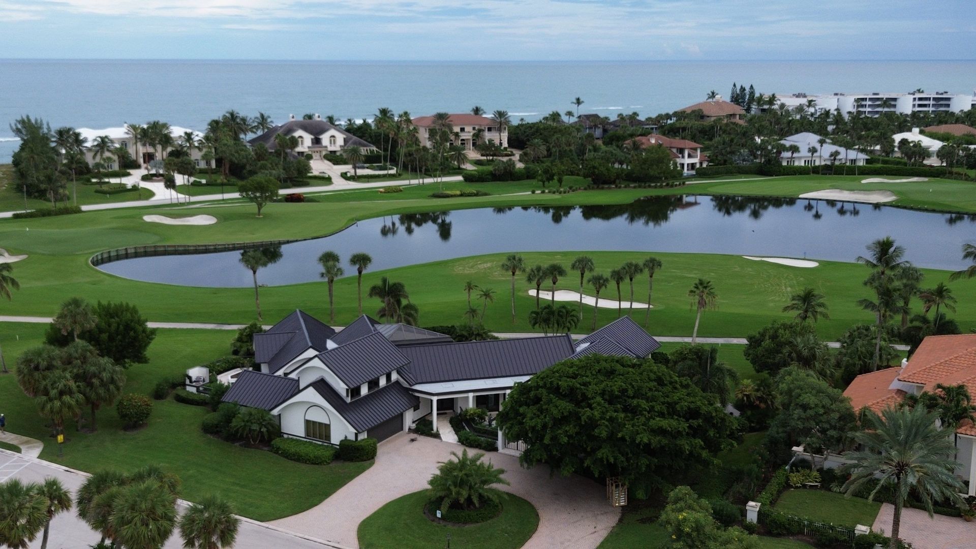 Aerial view of a luxury home with a long driveway, green lawn, and adjacent golf course and ocean.