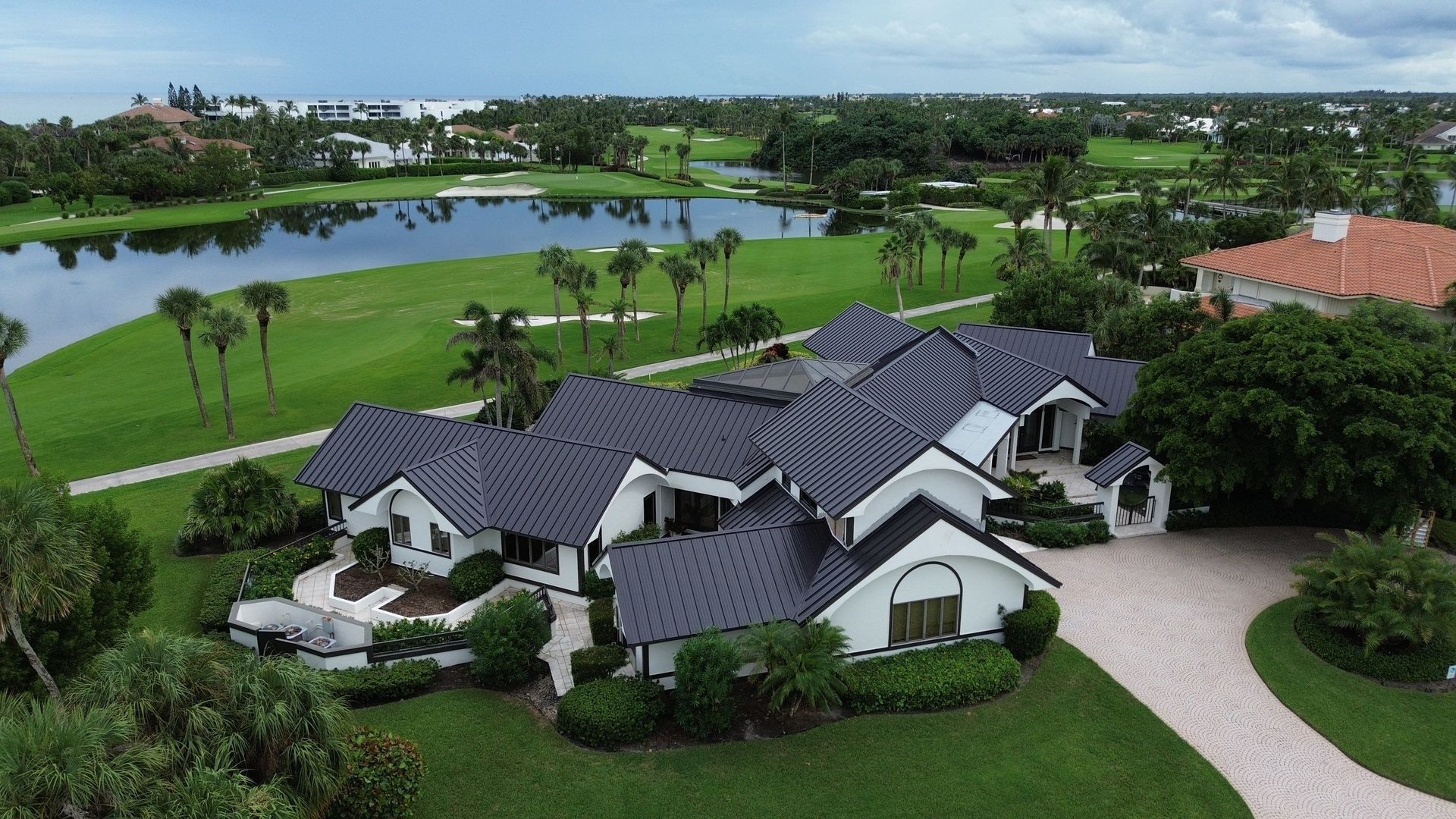 Large white house with a dark roof next to a golf course, lake, and lush green vegetation. A long curved driveway leads to the home.
