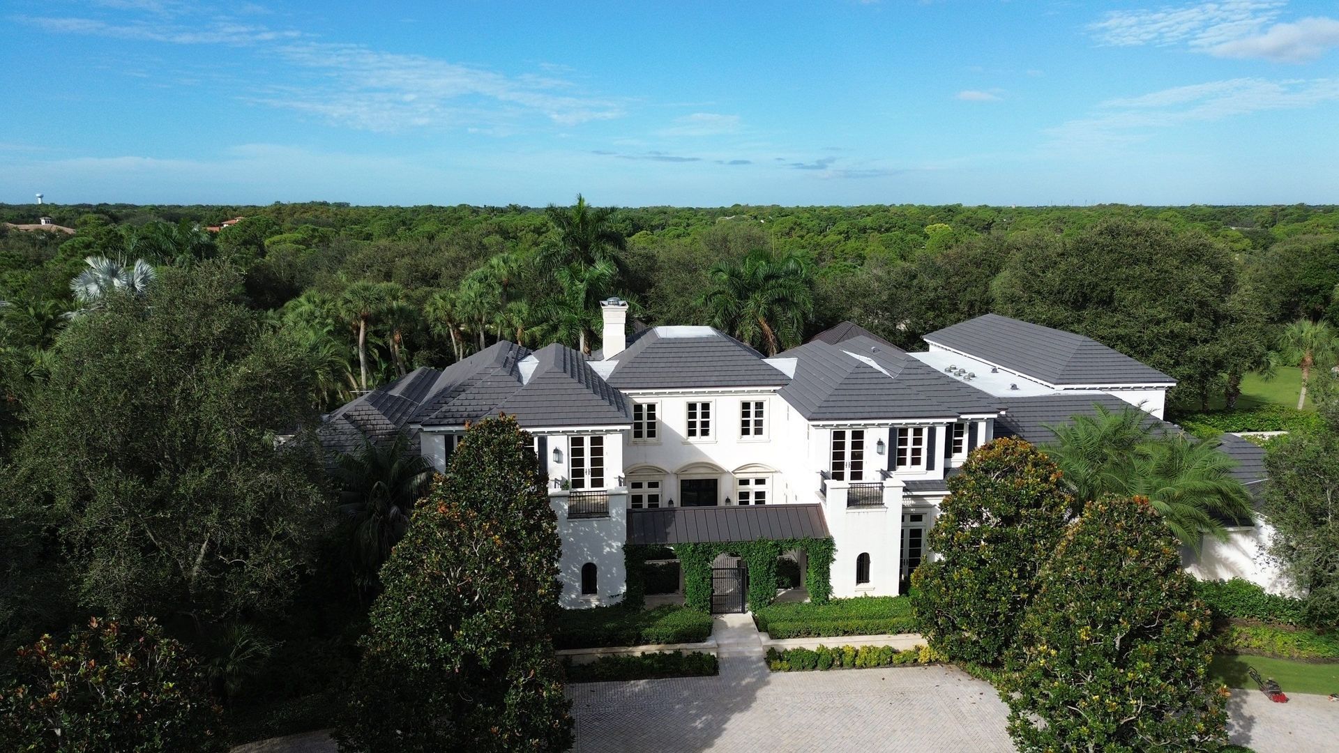 Large white mansion with dark roof surrounded by lush green trees under a bright blue sky.