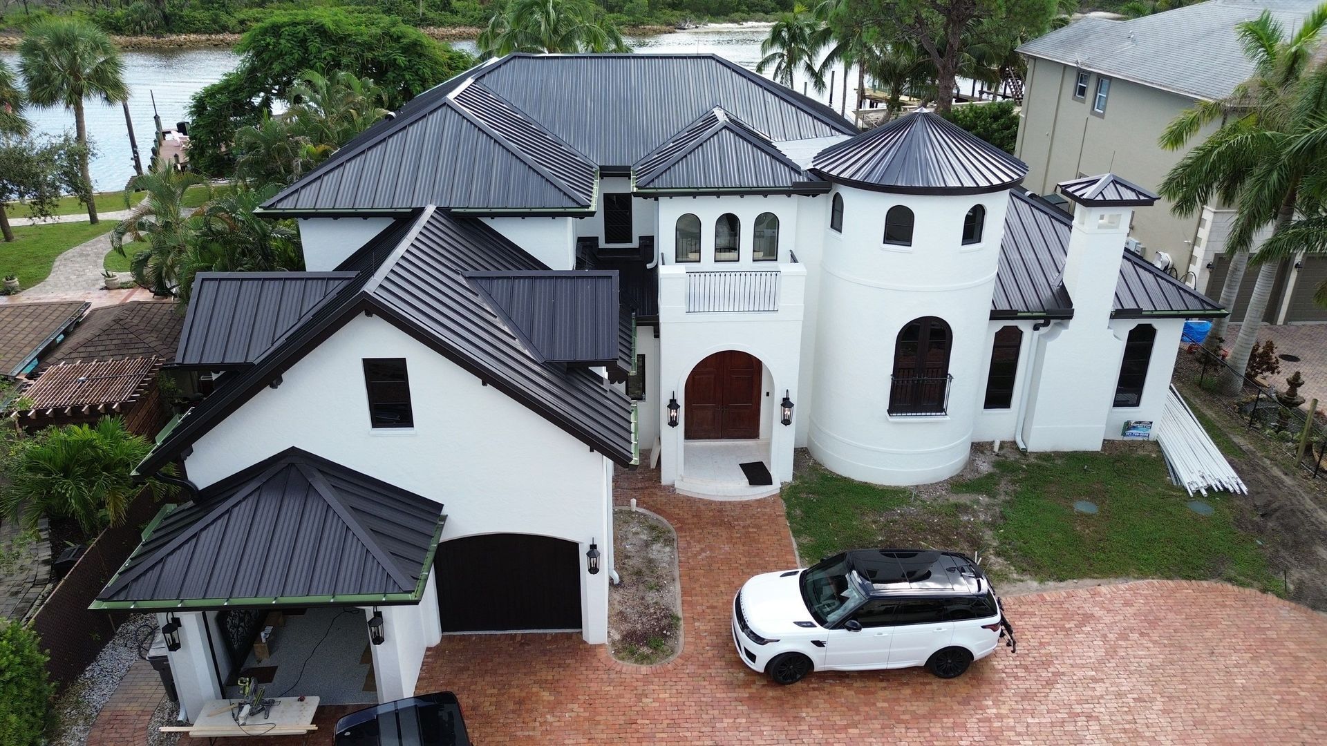 White stucco house with black roof and a circular turret, set on a red-brick driveway with a white SUV parked in front; next to a river with palm trees.