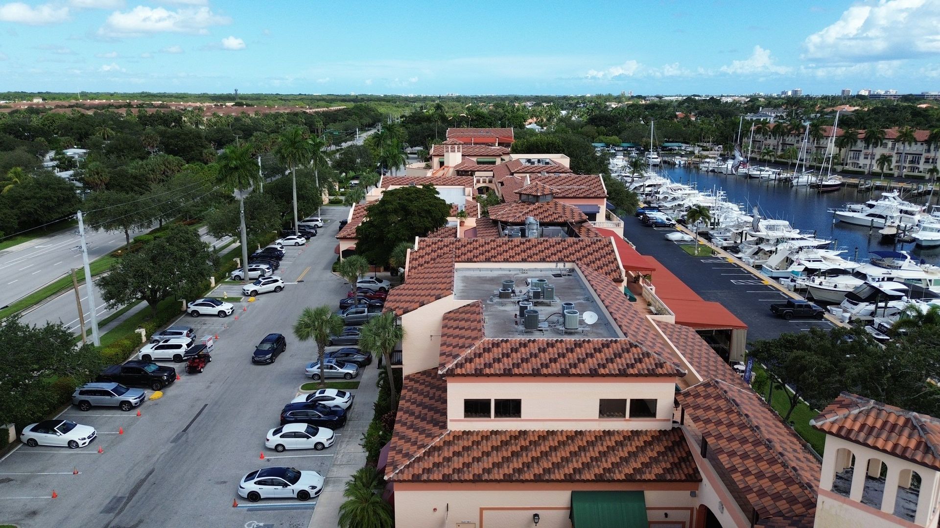 Aerial view of a long, red-tiled building with cars parked along a street and a marina with boats visible in the background.