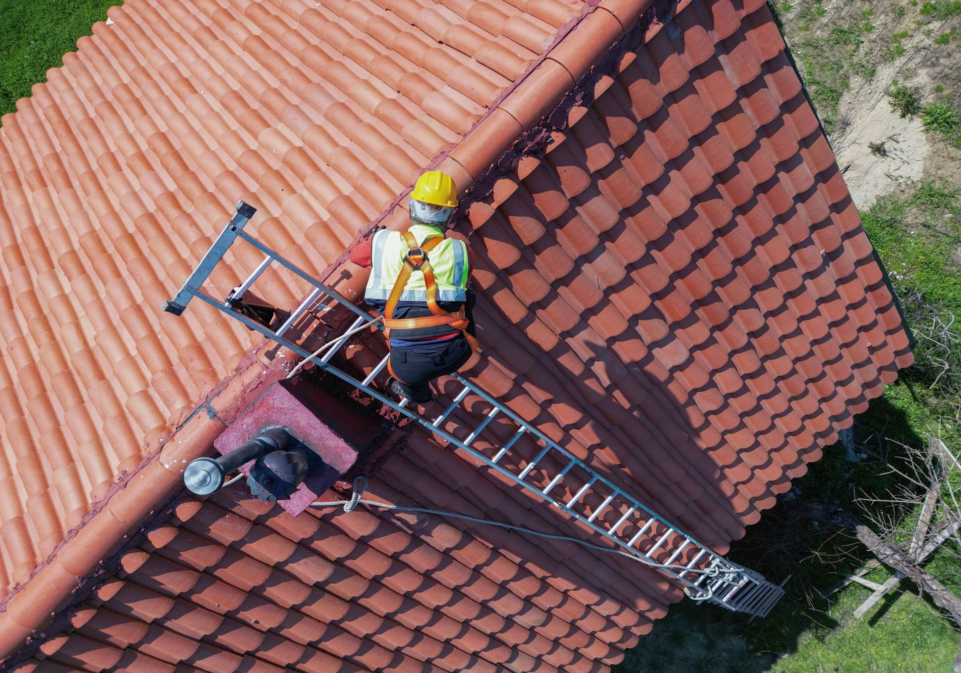 A male tile roofing contractor with safety gear is working on a steep red tile roof in daylight.