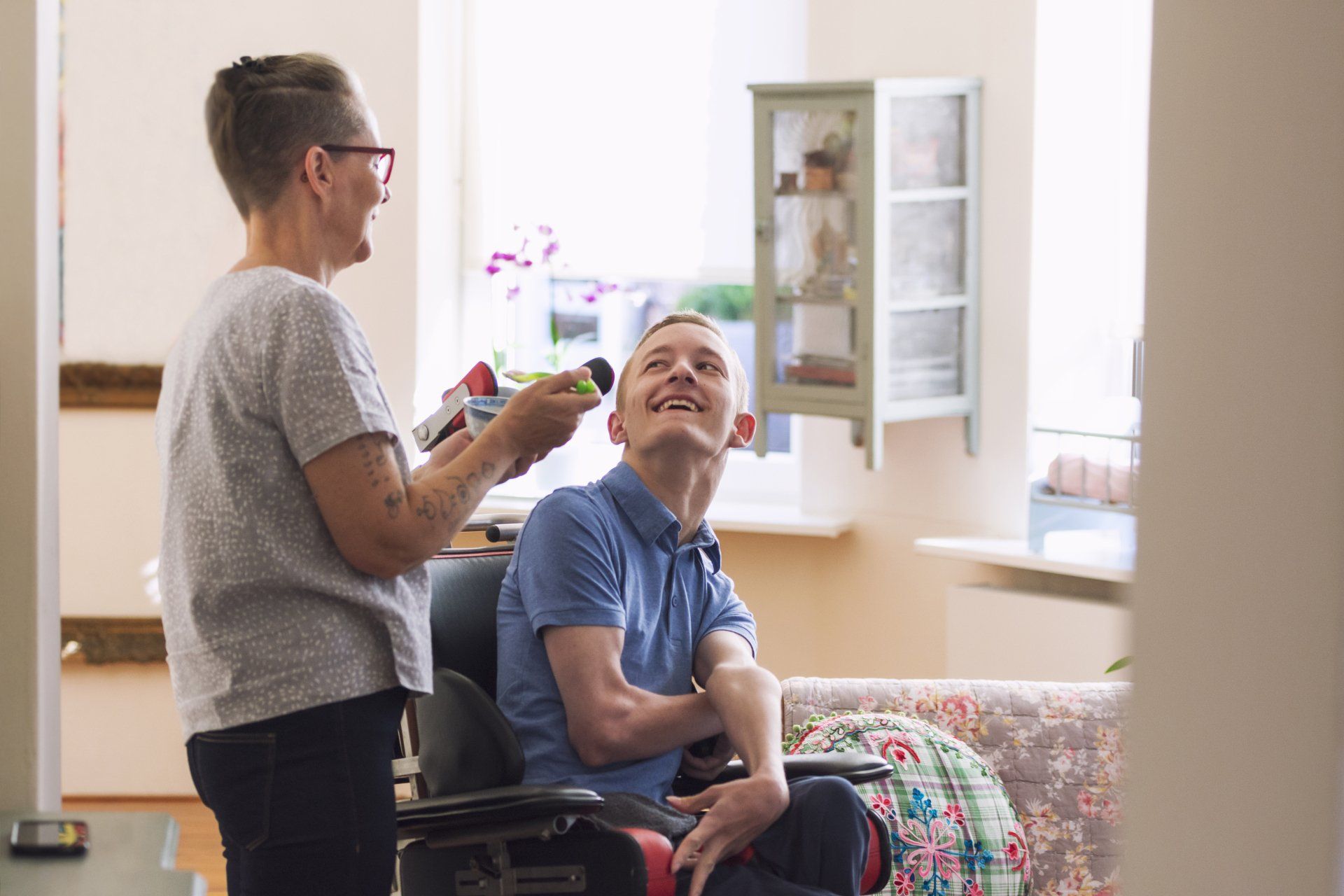 Woman brushes man's hair, both smiling, in a bright living room. Man is in a wheelchair.