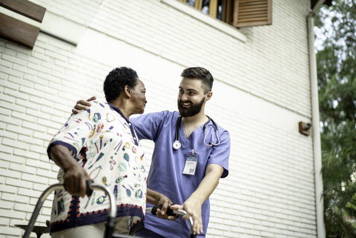 Nurse assists older Black woman with walker; smiles, outside a building.