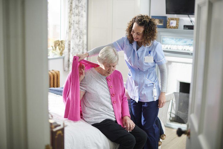 Caregiver helping elderly woman put on pink cardigan in a bedroom.
