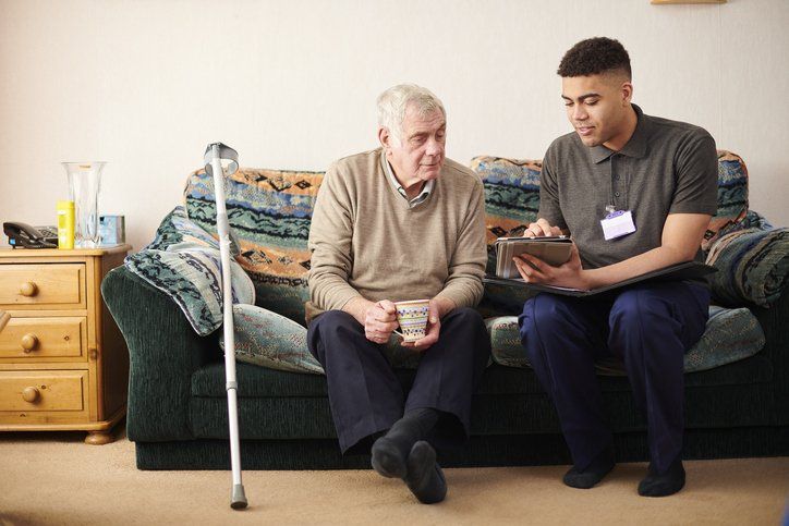 Man in beige sweater sits with a caregiver, both looking at a tablet on a couch; cane nearby.