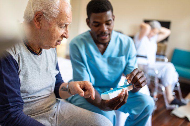 A healthcare worker showing an older man a pill organizer; another patient sits in the background.