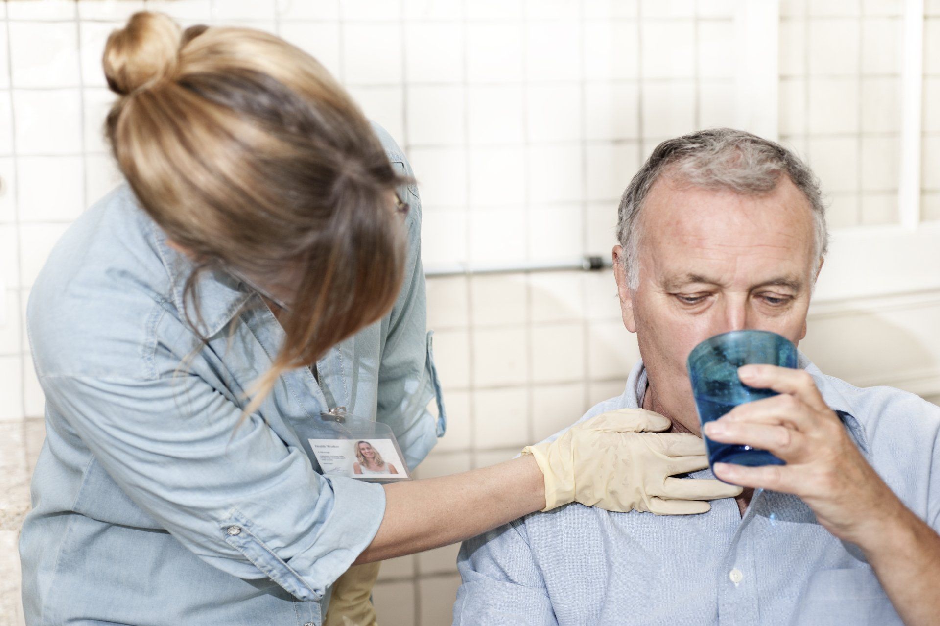 Woman in scrubs assists an older man drinking water, hand on his throat, in a clinical setting.