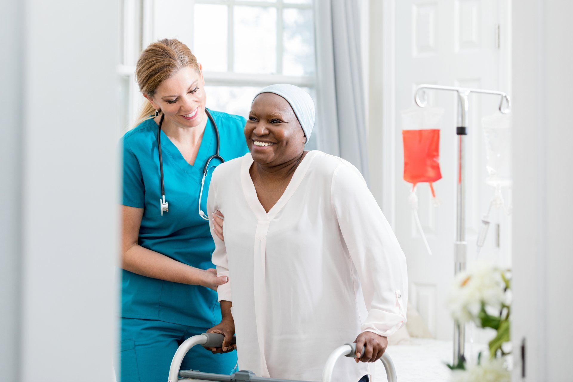 Nurse assists a smiling Black woman using a walker. White room with IV stand, flowers.