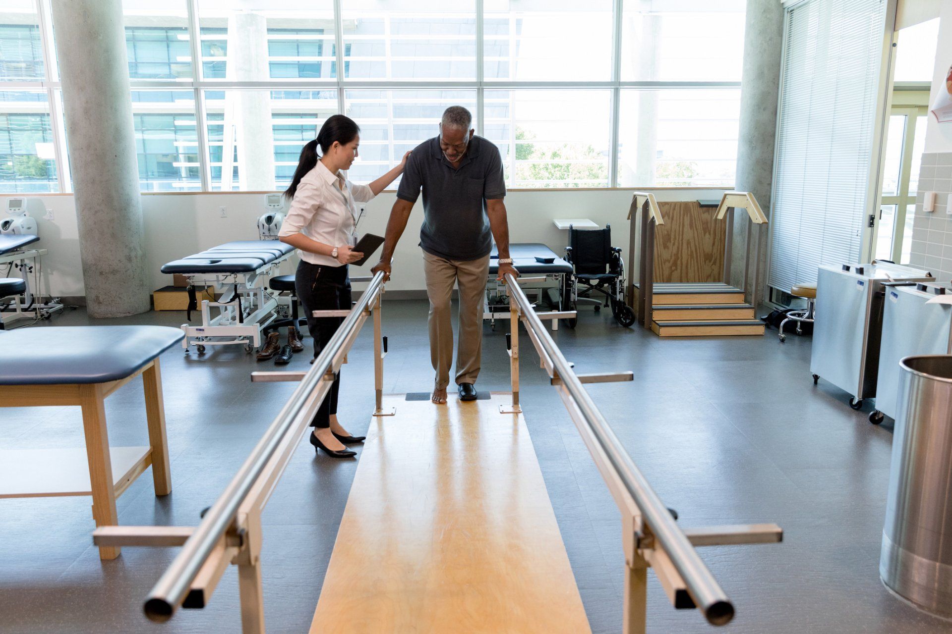A physical therapist helps a Black man walk between parallel bars in a rehab center.