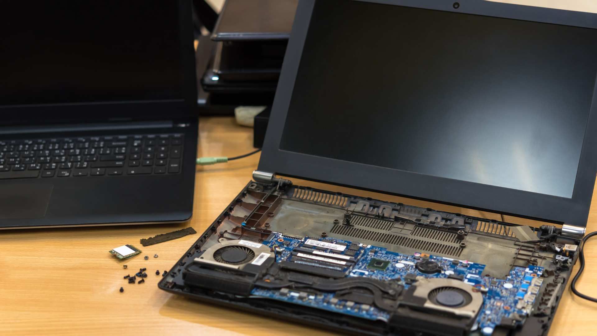 Open laptop beside a disassembled laptop with exposed internal components on a wooden desk.