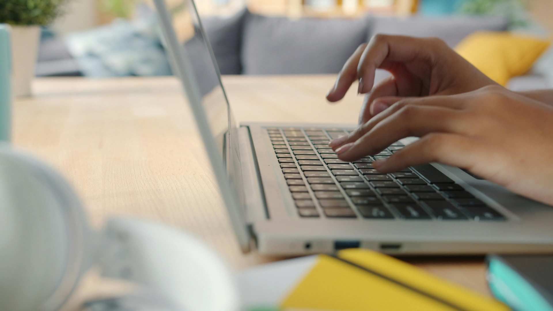 Hands typing on a laptop at a desk with a blurred notebook and yellow stationery nearby