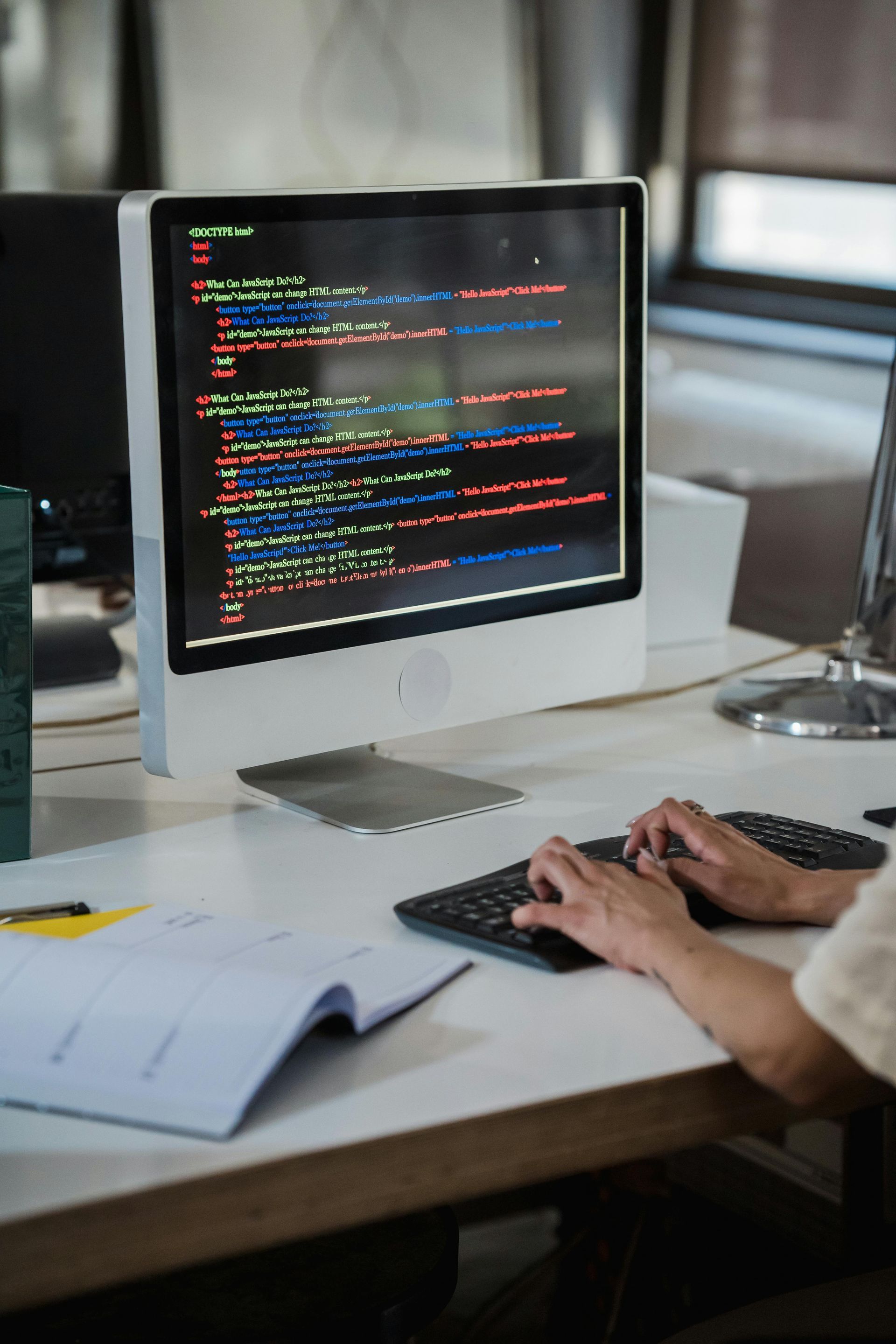 Person typing at a desktop computer with colorful code on the screen in a desk workspace