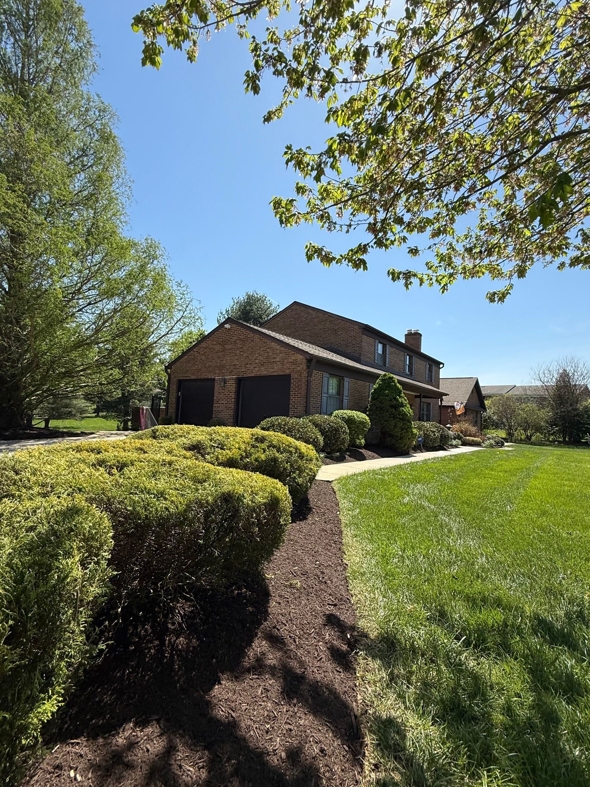 Brick house with brown roof beside trimmed hedges and a green lawn under a clear blue sky