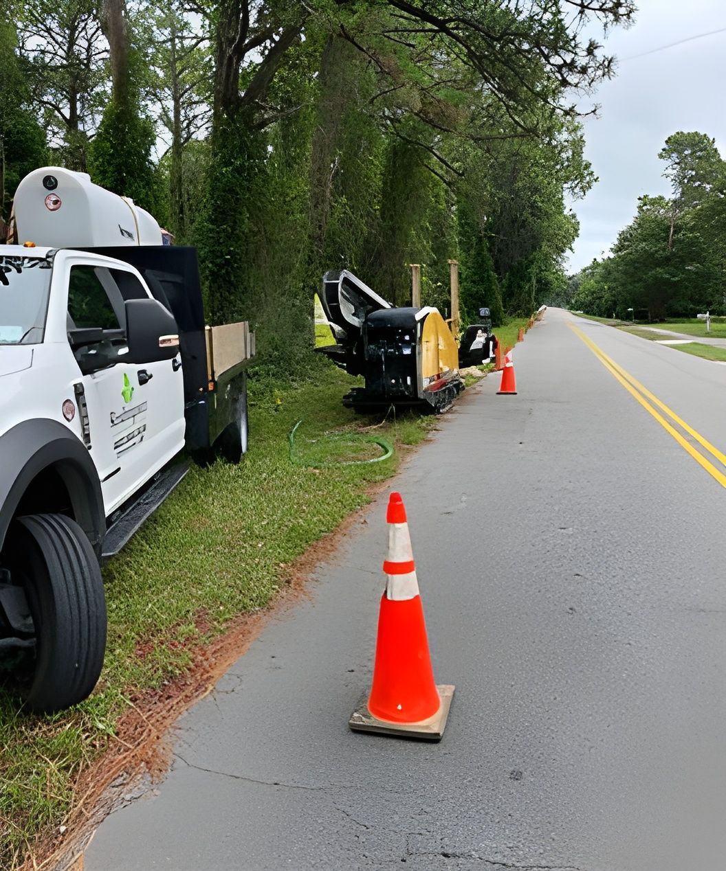Roadside work zone with orange cones, trucks, and equipment along a tree-lined street.